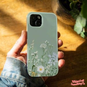 May include: A mint green phone case featuring a floral design with white and yellow wildflowers. The case has a glossy finish and is held in a person's hand. The phone case has a dual camera system. The text "Whimsical Something Co" is in the bottom right corner.