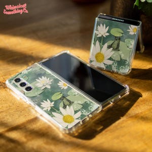 May include: A clear phone case with a green, white, and yellow floral design featuring a frog sitting on a lily pad. The case is designed for a Samsung foldable phone.