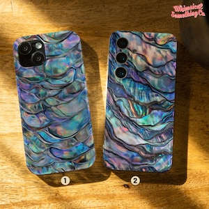 Op de afbeelding: Twee iriserende telefoonhoesjes met een wervelend, getextureerd ontwerp in tinten blauw, paars en groen. De hoesjes hebben camera-uitsparingen en zijn genummerd 1 en 2. De hoesjes liggen op een houten oppervlak.
