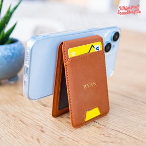 May include: A brown leather phone wallet with a card inserted, personalized with the name "RYAN". The wallet is attached to a clear phone case. The background includes a plant and the text "Whimsical Something Co."