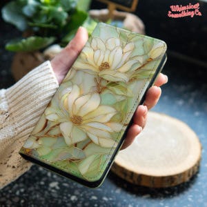 May include: A rectangular phone case featuring a stained glass style design of white magnolia flowers with gold outlines against a pale green background. The text Whimsical Something Co is printed in pink in the upper right corner of the case.
