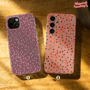 May include: Two phone cases with different patterns. The first case is a light pink with white polka dots. The second case is a light pink with a green leaf pattern. Both cases have a camera cutout.