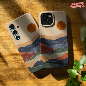 Puede incluir: Dos fundas para tel&eacute;fono con un fondo beige y un dise&ntilde;o de paisaje. El dise&ntilde;o presenta una cordillera de estilo acuarela con acentos azules, verdes, rojos y dorados. La parte superior de la funda tiene un c&iacute;rculo de color naranja similar a una puesta de sol.