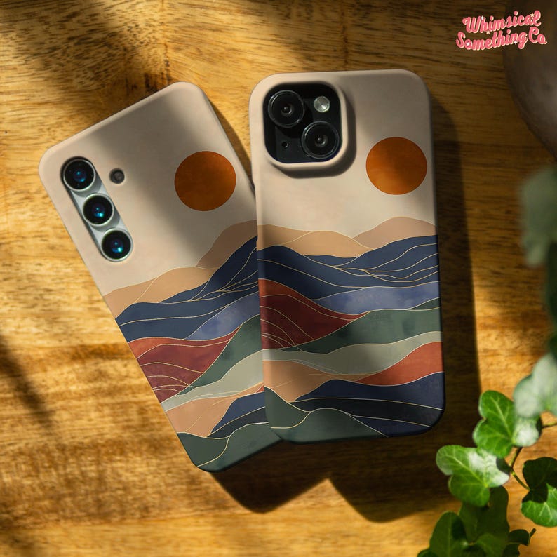 Puede incluir: Dos fundas para tel&eacute;fono con un fondo beige y un dise&ntilde;o de paisaje. El dise&ntilde;o presenta una cordillera de estilo acuarela con acentos azules, verdes, rojos y dorados. La parte superior de la funda tiene un c&iacute;rculo de color naranja similar a una puesta de sol.