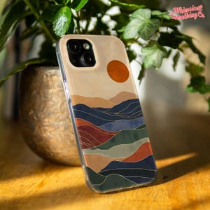 May include: A clear phone case with a landscape design. The design features a sunset and layered hills in beige, blue, red, and green, with gold outlines. The text "Whimsical Something Co." is visible in the upper right corner of the case.