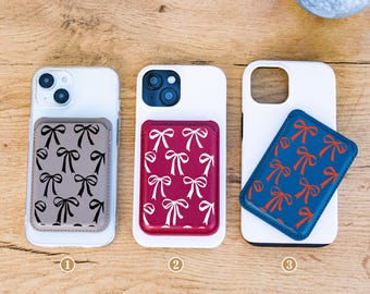 May include: Three phone cases with attached cardholders. The cardholders feature bow patterns in black on gray, white on burgundy, and orange on blue. The cases are on a wooden surface. The top left corner has the text "Whimsical Something Co."
