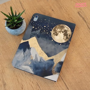 May include: An iPad case featuring a celestial design with a full moon, gold accents, and a mountain range. The case is navy blue with white and gold details. A small succulent plant in a gray pot sits to the left of the case.