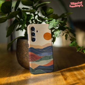 Könnte beinhalten: Eine Handyhülle mit einem farbenfrohen abstrakten Landschaftsdesign. Die Hülle zeigt einen Sonnenuntergang mit einer großen orangen Sonne, blauen und grünen Bergen und einem braunen und orangen Vordergrund. Die Hülle hat einen Kameraausschnitt mit drei Objektiven.