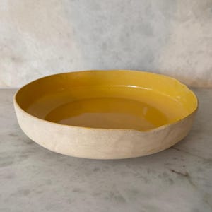 Puede incluir: Un cuenco redondo y poco profundo con un interior amarillo brillante y un exterior color crema. El cuenco tiene un diseño simple y minimalista y parece estar hecho de cerámica o un material similar. El cuenco está sobre una superficie de mármol.