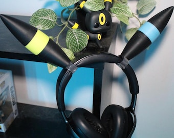May include: Black headphones with large earcups and a headband with attached black and yellow accents. A black Umbreon figure with yellow accents is visible in the background. The headphones are resting on a black surface.