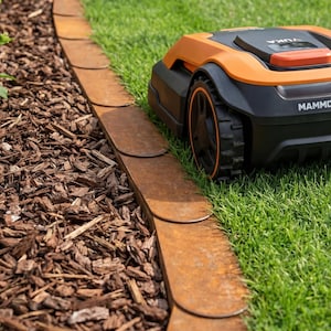 K&ouml;nnte beinhalten: Ein orange-schwarzer Roboter-Rasenm&auml;her auf einem gr&uuml;nen Rasen neben einer rostigen Metall-Gartenbegrenzung. Die Begrenzung trennt das Gras von einem Beet aus braunem Mulch. Auf dem Rasenm&auml;her steht "MAMMO".