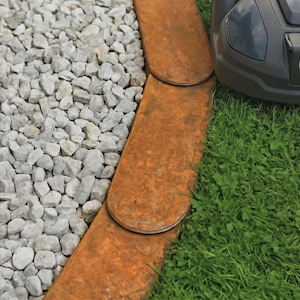 May include: A close-up view of a garden bed border made of rusty metal edging. The edging separates a bed of gray gravel from a patch of green grass. A robotic lawnmower is visible on the right side of the image.