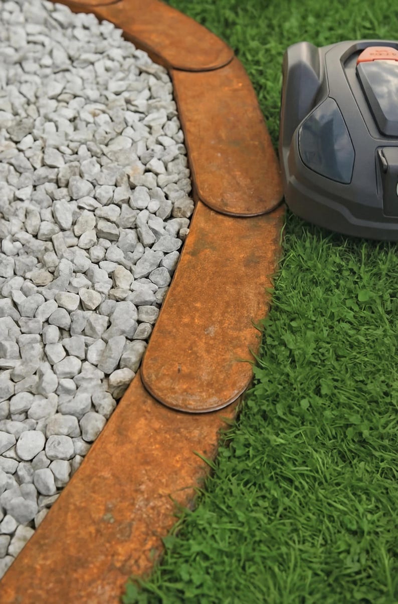 K&ouml;nnte beinhalten: Nahaufnahme einer Gartenbegrenzung aus rostigem Metall. Sie trennt grauen Kies von gr&uuml;nem Rasen. Auf der rechten Seite ist ein Roboter-Rasenm&auml;her zu sehen. Die Metallplatten sind gebogen und &uuml;berlappen sich.