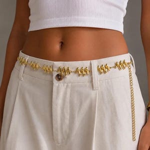 May include: A gold-toned chain belt featuring a series of repeating, twisted, zigzag-shaped metal links. The belt is worn around the waist over light-colored trousers, with a single dangling chain segment extending from the side of the waistline.