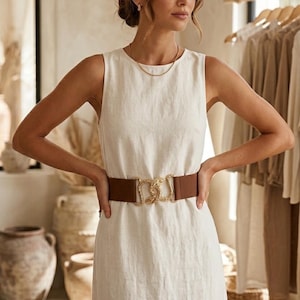 Könnte beinhalten: Ein ärmelloses, cremefarbenes Leinenkleid mit einem braunen elastischen Gürtel und einer goldfarbenen Schnalle. Das knielange Kleid hat ein schlichtes, elegantes Design. Der Gürtel betont die Taille. Im Hintergrund Kleiderstangen und dekorative Vasen.