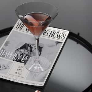 Könnte beinhalten: Ein Martini-Glas gefüllt mit einem rosa Getränk steht auf einer Hochzeitsanzeige mit der Überschrift "Happily Ever After" und dem Datum "June 19th 2026".