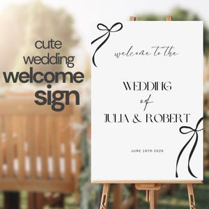 Könnte beinhalten: Weißes Hochzeits-Willkommens-Schild mit schwarzem Kalligraphie-Text, der "Willkommen zur Hochzeit von Julia & Robert" und "19. Juni 2026" lautet. Das Schild ist mit schwarzen Schleifen verziert.