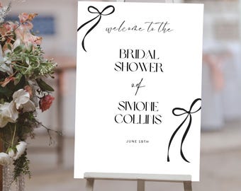 Pode incluir: Um cartaz de boas-vindas branco com caligrafia preta que diz "Bem-vindo ao Bridal Shower de Simone Collins em 19 de junho". O cartaz tem dois laços de fita pretos de cada lado.
