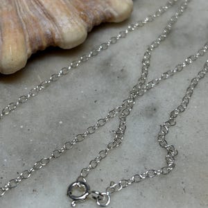 May include: A delicate silver chain necklace with a lobster clasp. The chain is made of small, interconnected oval links. The necklace is displayed on a light-coloured, textured surface, with a seashell in the background.