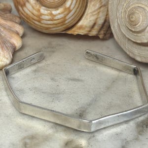 May include: A silver cuff bracelet with a hexagonal shape and a smooth, polished finish. The metal band features an open design, allowing it to be slipped onto the wrist. A small 925 hallmark is stamped on the inner surface of the silver band.