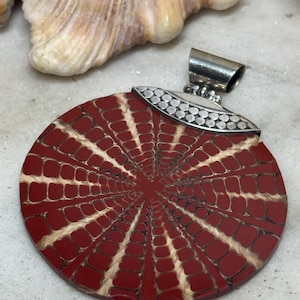 Puede incluir: Un colgante circular con una piedra de color rojo intenso que presenta un patrón radiante similar a una telaraña con líneas de color crema. La parte superior tiene una anilla de metal plateado curvada y texturizada con pequeños puntos.