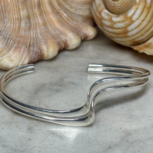 May include: A polished silver cuff bracelet featuring a double-band design that curves into a wave-like shape. The metal has a smooth, reflective surface and an open back, showcasing a clean, metallic aesthetic with no additional embellishments.