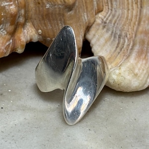 Op de afbeelding: Een gepolijste zilveren ring met een glad, doorlopend wikkelontwerp met twee puntige, overlappende uiteinden. Het metalen oppervlak is reflecterend en vertoont subtiele gebruikssporen, wat zorgt voor een strakke, vloeiende vorm rond de vinger.