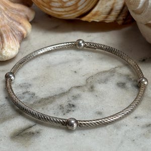 May include: A silver bangle bracelet with a textured, twisted rope design. The bracelet has four small, round, silver beads evenly spaced. The jewellery is displayed on a marble surface.