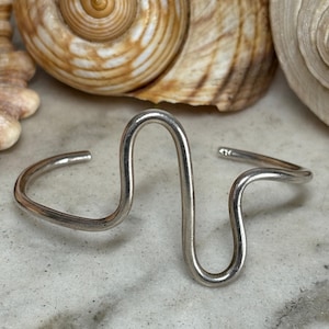 May include: A silver-toned metal cuff bracelet featuring a continuous, fluid, wave-like design that rises into a prominent central peak and a smaller secondary curve. The bracelet has an open-ended construction with smooth, rounded edges.