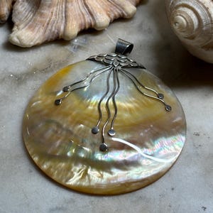May include: A large, iridescent pendant with a natural shell design. The pendant features a round, cream-coloured shell with yellow and silver tones. Delicate silver wire detailing extends from the top, adding a unique touch to this handcrafted jewellery piece.