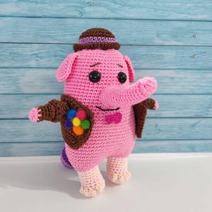 May include: A pink crocheted elephant plush toy wearing a brown hat and a brown jacket with a colorful paint palette. The elephant has a long pink trunk and black eyes.