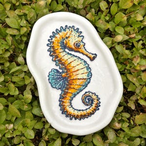 May include: A white, irregularly shaped ceramic dish featuring a detailed seahorse design. The seahorse is rendered in a mosaic style, with shades of orange, yellow, and blue. The dish is set against a backdrop of green foliage.