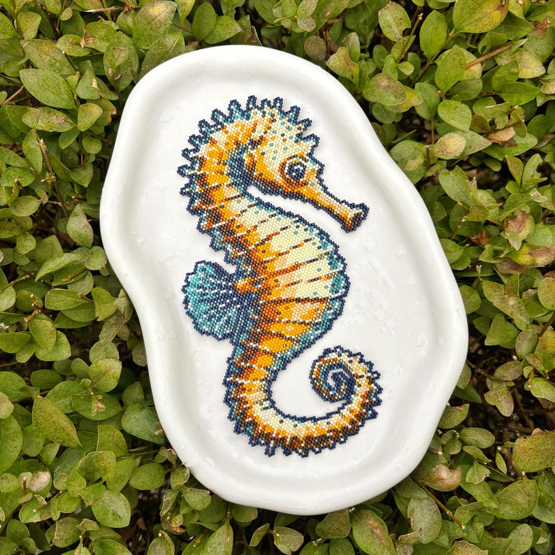 May include: A white, irregularly shaped ceramic dish featuring a detailed seahorse design. The seahorse is rendered in a mosaic style, with shades of orange, yellow, and blue. The dish is set against a backdrop of green foliage.