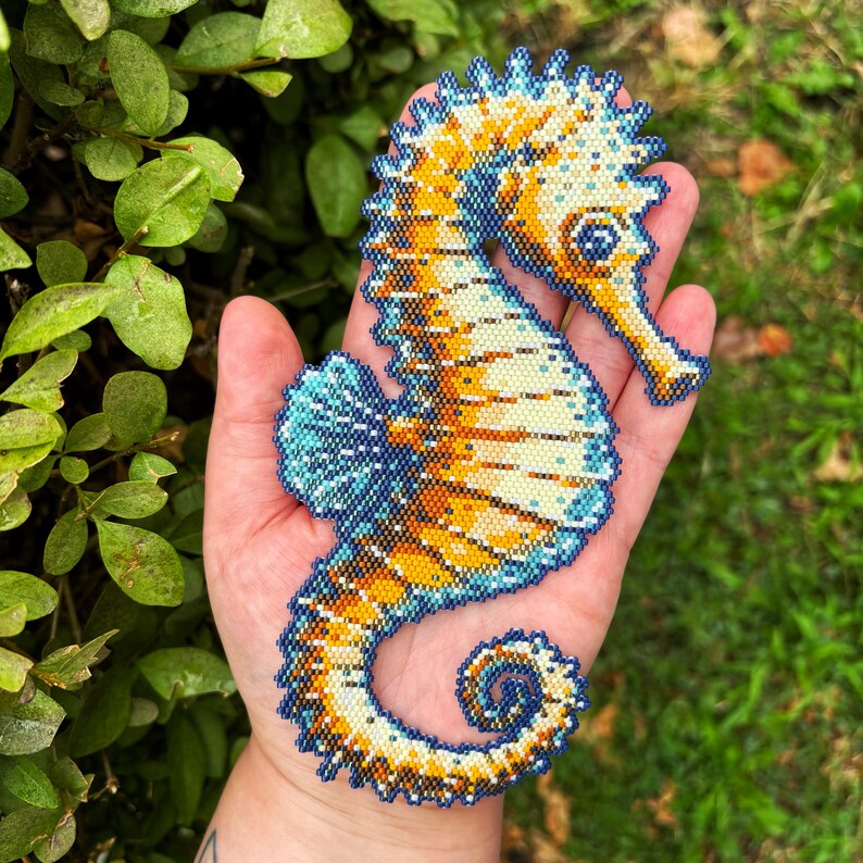 May include: A beaded seahorse in shades of blue, yellow, and white. The seahorse is intricately detailed with a textured surface. The seahorse is held in a hand against a background of green foliage and grass.