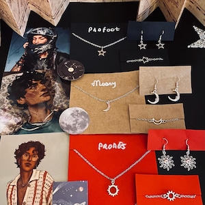 Könnte beinhalten: Eine Sammlung von silbernen Halsketten, Ohrringen und Armbändern mit Anhängern zum Thema Sterne und Himmelskörper. Der Schmuck wird auf schwarzen und roten Karten präsentiert, auf denen die Wörter "PAOFOOT", "Moony" und "PRONES" stehen. Das Bild enthält auch Illustrationen.