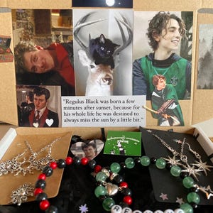 May include: A decorative box featuring images and jewelry. The box contains photos of a person, a black cat on a deer, and jewelry. The jewelry includes beaded bracelets with black, red, and green beads, and necklaces with star pendants.
