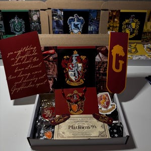 Könnte beinhalten: Eine Box mit Harry-Potter-Merchandise, darunter ein rotes Lesezeichen im Gryffindor-Design, eine Karte mit dem Gryffindor-Wappen und ein Ticket für Gleis 9 3/4. Die Box enthält auch andere Artikel im Gryffindor-Design.