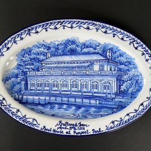 May include: A white oval ceramic platter with a blue and white design of a building with a lake in front of it. The text "Brittany & Sam April 5th, 2024 Boat house at Prospect Park" is written in blue on the bottom of the platter.