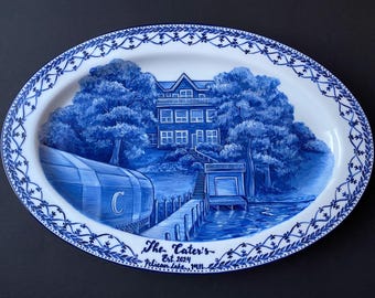 May include: Oval ceramic platter with a blue and white lakeside scene. The design features a house, trees, a dock, and a boathouse. The platter has a decorative border and text that reads "The Cater's Est. 1924 Pelican Lake, MN."