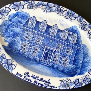 Pode incluir: Um prato oval azul e branco pintado à mão com uma borda floral. O centro do prato apresenta uma ilustração azul e branca de uma casa com uma grande árvore na frente. O texto "The Hall Family 3605 River Ferry Drive" está escrito em azul cursivo na parte inferior do prato.