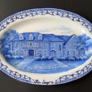May include: A blue and white oval ceramic platter with a hand-painted design of a house. The house is a two-storey brick home with white trim and a blue roof. The platter has a blue and white border with a floral design. The text "The Longo's" is written in cursive at the bottom of the platter.