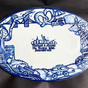 Può includere: Piatto ovale in ceramica bianca con un disegno blu dipinto a mano raffigurante i monumenti di New York City. Include il ponte di Brooklyn, la Statua della Libertà e un carretto di hot dog. Il testo recita "Michael & Lauren The Bowery Hotel December 2025."