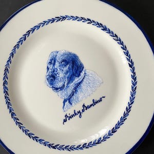 Puede incluir: Plato de cerámica blanco con borde azul e ilustración azul detallada de la cara de un perro. El plato presenta un patrón decorativo de hojas azules alrededor del borde y la inscripción "Binky Bambino".