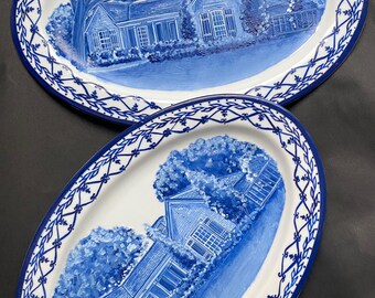 May include: Two blue and white oval ceramic plates with a hand-painted design of a house and trees. The plates have a blue and white border with a vine pattern. The bottom plate has the text "The Venner's Est. 1957" written in blue.