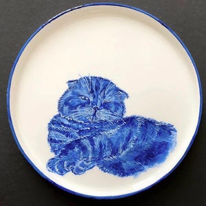 Pode incluir: Um prato de cerâmica branco com uma ilustração de um gato azul pintada à mão. O gato está deitado com os olhos fechados.
