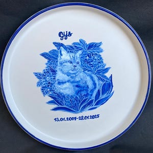 Peut inclure: Assiette en céramique blanche avec un bord bleu, ornée d'une illustration de chat bleu et blanc peinte à la main, entourée d'éléments floraux. L'assiette porte le nom "Gijs" et les dates 13.01.2007-28.01.2025.