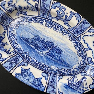 Könnte beinhalten: Ovale Keramikplatte mit blau-weißem Design. Die Platte zeigt eine zentrale Landschaftsszene, umgeben von floralen und nautischen Motiven. Enthält Bilder von Katzen, einem Segelboot und den Text "Martha's Vineyard".
