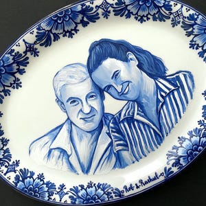Puede incluir: Un plato ovalado de cerámica blanca con un diseño floral azul y blanco. El centro del plato presenta un retrato pintado de una pareja en azul y blanco. La pareja sonríe y la mujer apoya la cabeza en el hombro del hombre. El plato está firmado "Rob & Rachael" en azul.