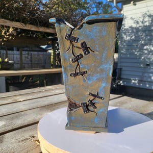 May include: A rectangular ceramic vase with a distressed blue and gold finish. The vase features decorative black accents resembling stitches and a partially broken edge, adding a unique, handcrafted aesthetic. The vase is displayed on a white surface.