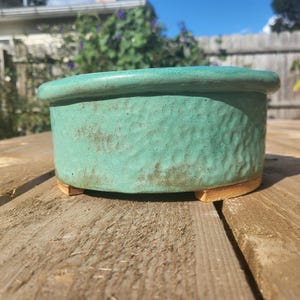 Puede incluir: Una maceta de cerámica turquesa con superficie texturizada y borde redondeado. La maceta se apoya sobre tres pequeños pies rectangulares. El exterior tiene un aspecto moteado con toques marrones.